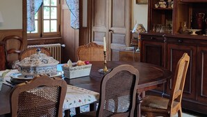 A dining room with a wooden table, four chairs, a chandelier, a patterned rug, and a bookshelf with books and decorative items. - Chateau de la Mothe (Saint-Sulpice-en-Pareds)
