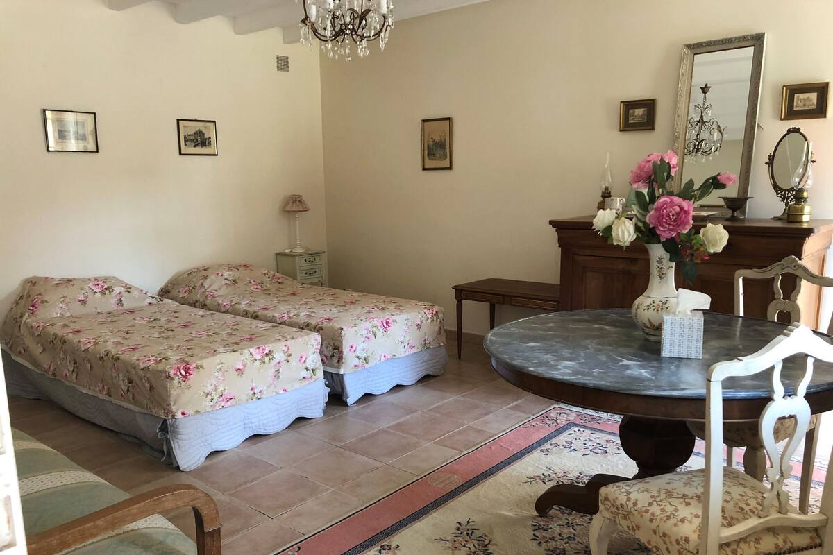 A bedroom with two beds, a dining table, a chair, a vase with flowers, and a mirror.