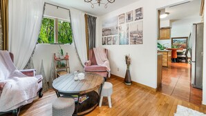 Un salon chaleureux avec une table ronde en bois, deux fauteuils roses et un petit tabouret blanc. Une grande fenêtre, ornée de rideaux, offre une vue sur la verdure à l’extérieur.