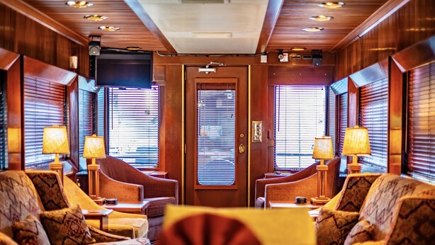A hotel room with a wooden interior, a large television, and two windows with blinds. There are two armchairs and a table with lamps. - Rail Hotel  (Galveston)