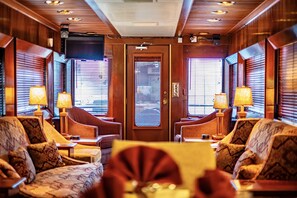 A hotel room with a wooden interior, a large television, and two windows with blinds. There are two armchairs and a table with lamps. - Rail Hotel (Galveston)