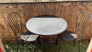 Mesa redonda de metal com quatro cadeiras, colocada numa área gramada com uma cerca de bambu ao fundo.
