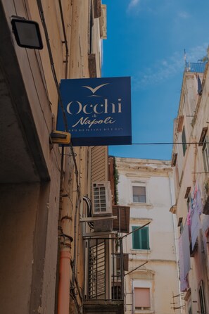 Une rue étroite, bordée de bâtiments des deux côtés, avec une enseigne bleue indiquant « Oecchi Napoli », ainsi que des tuyaux et des balcons apparents.