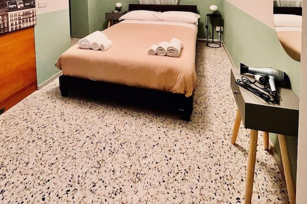 A hotel room with a bed, a nightstand, a mirror, a window with curtains, and a wall with a cityscape painting.