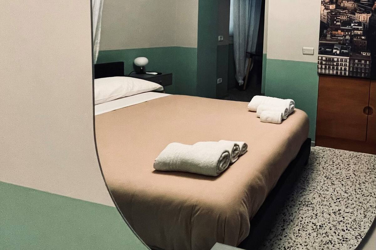 A hotel room with a bed, a nightstand, a mirror, a painting, and a view of a cityscape.