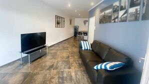 A modern living room with a flat-screen TV, a black leather sofa with blue and white striped pillows, and a dining area in the background.
