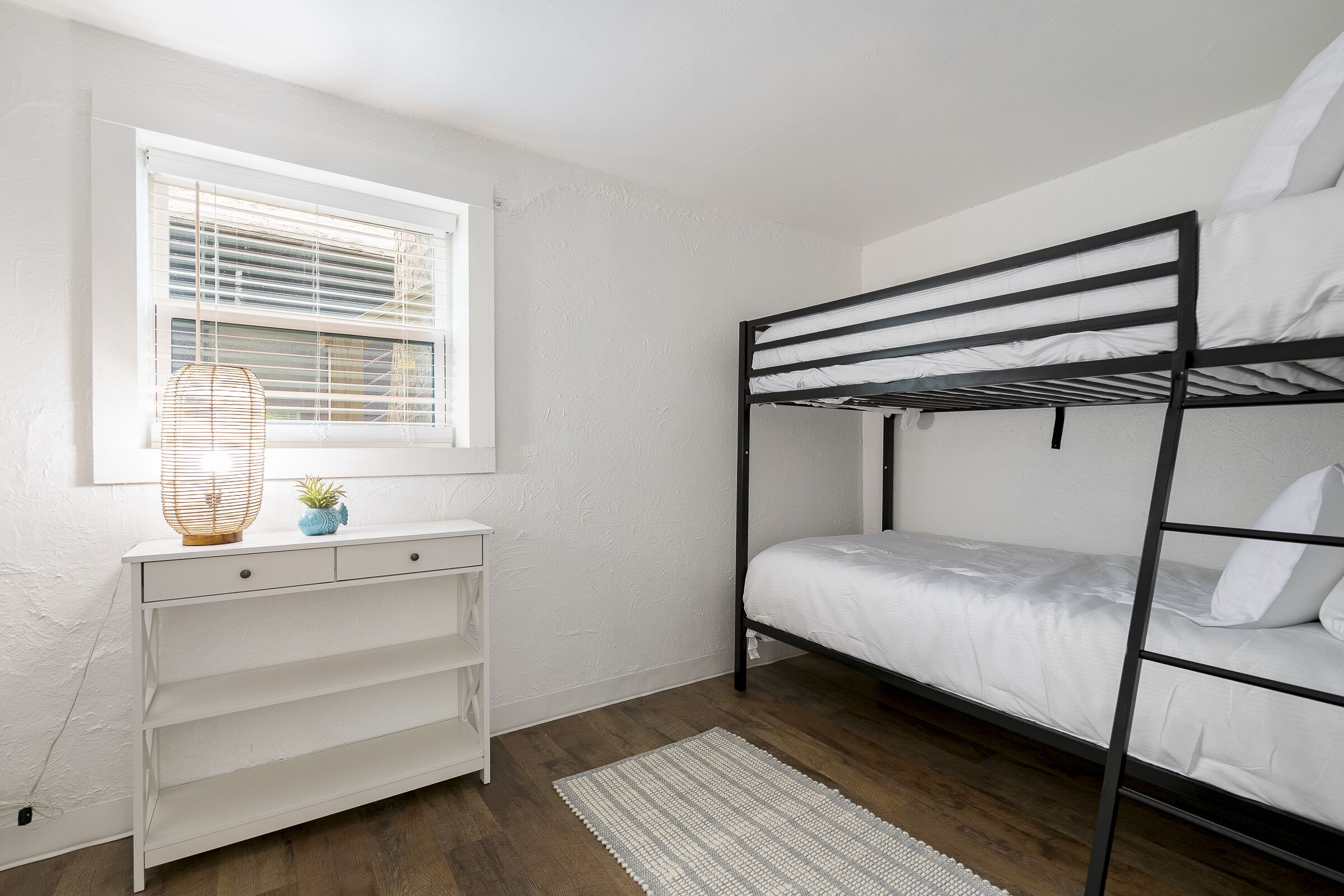 A bunk bed with white bedding, a white desk with drawers, a small plant, and a window with blinds.