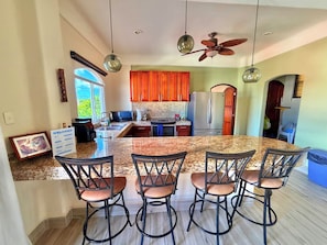 A modern kitchen with granite countertops, a central island with bar stools, wooden cabinets, and a stainless steel refrigerator.