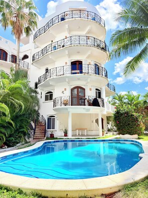 A multi-story building with balconies, a swimming pool, and palm trees.