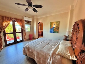 A bedroom with a bed, a ceiling fan, an air conditioning unit, a painting, and a view through a window.