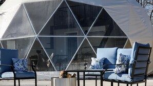 A geodesic dome structure with a transparent section, surrounded by outdoor seating with blue cushions and a metal table.