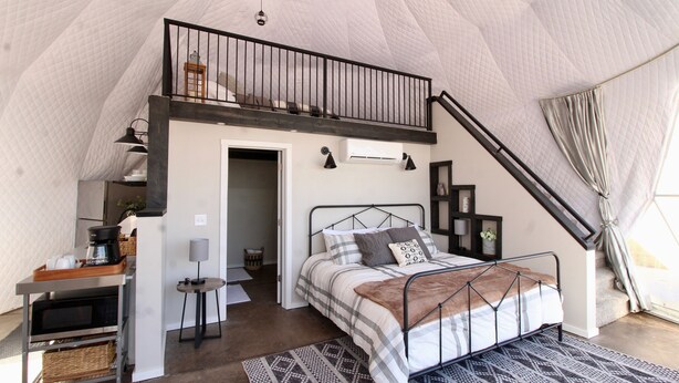 A bedroom with a loft bed, a desk with a coffee maker, and a view of the outdoors.
