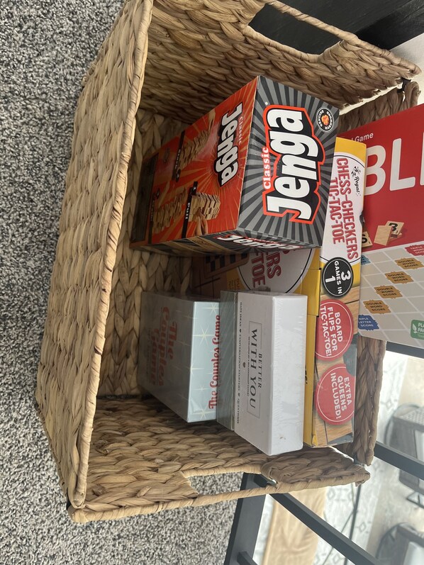 Una cesta tejida contiene varios juegos de mesa, incluyendo 'Jenga' y 'Ajedrez Damas'.