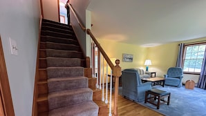 A staircase with wooden handrails leading to a living room with a blue armchair, a wooden coffee table, and a lamp.