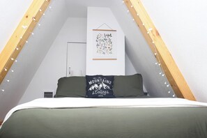 A bedroom with a sloped ceiling, a bed with a green bedspread, a pillow with a mountain design, a wall-mounted light fixture, and a framed botanical print. - A-Frame Cabin - 5 Mins to NP - Tilted Timbers (Ashford)