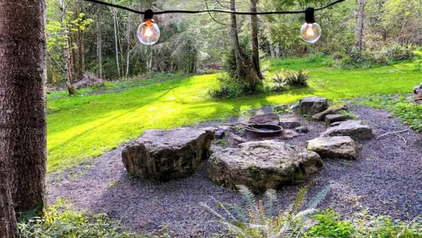 Un espace extérieur rustique doté d’une structure en bois, d’un foyer en pierre et d’une pelouse verdoyante entourée d’arbres.