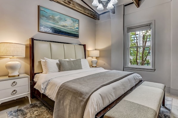 A bedroom with a large bed, two bedside tables with lamps, a window with blinds, and a painting on the wall.