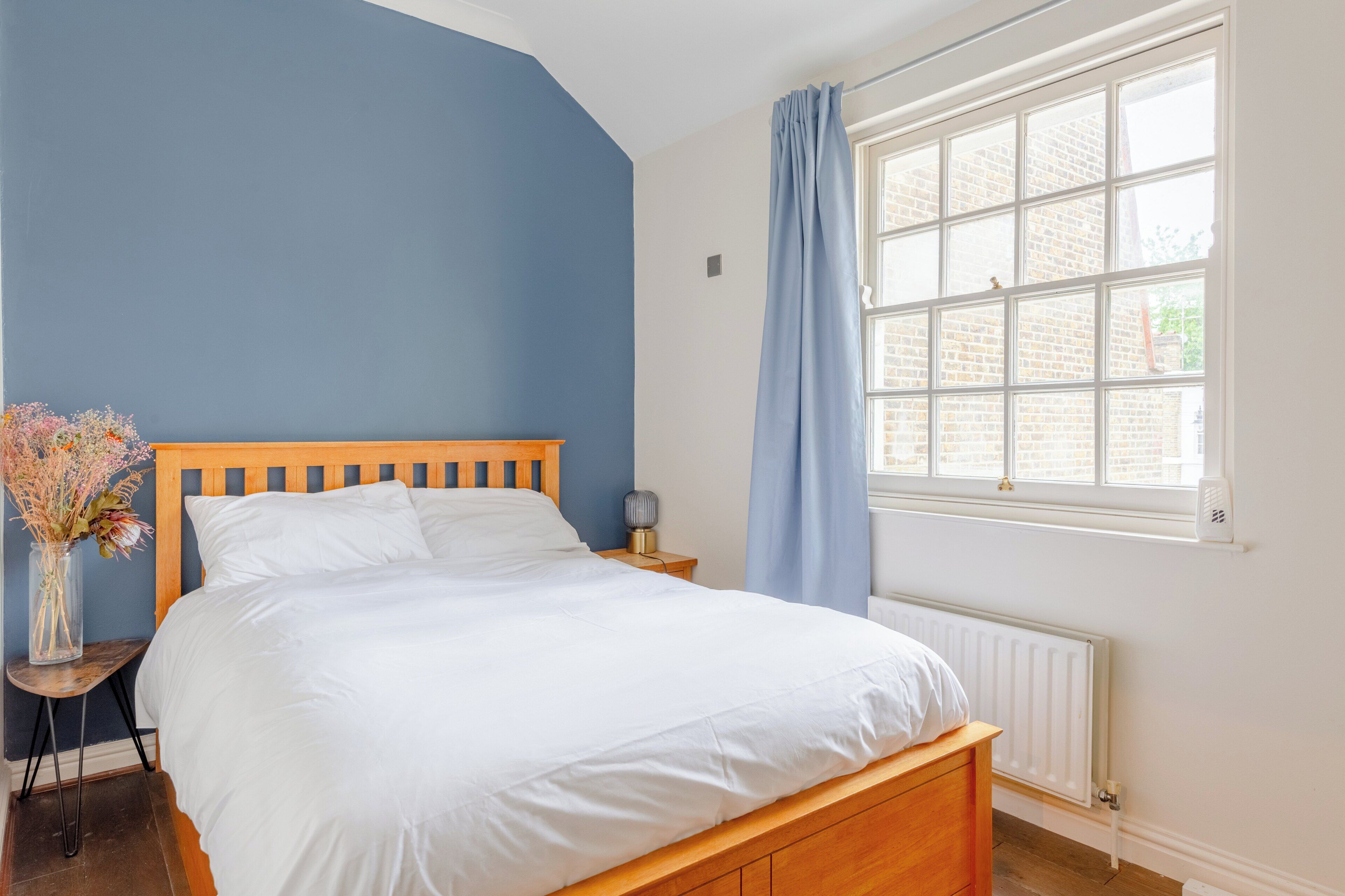 Een slaapkamer met een houten bed, een nachtkastje, een vaas met bloemen en een raam met gordijnen.