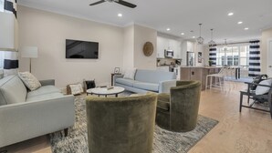 A modern living room with a sofa, armchairs, a coffee table, and a TV. A kitchen is visible in the background. - Downtown Greenville · 3BR/3.5BA · Private 2-Car Garage · Minutes to Main St (Greenville)