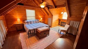 A cozy log cabin bedroom with a bed, bedside tables, a fireplace, and a chair. - Naked Creek Cabin (Page County)
