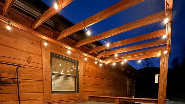 A wooden cabin with a hot tub under a wooden pergola, string lights, and a window with blinds. - Naked Creek Cabin (Page County)
