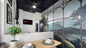 A modern dining area with a wooden table, white plates, and a glass of wine. A potted plant is on the left, and a mural of a landscape is on the wall. - The Neon Elm Hotel (San Diego)