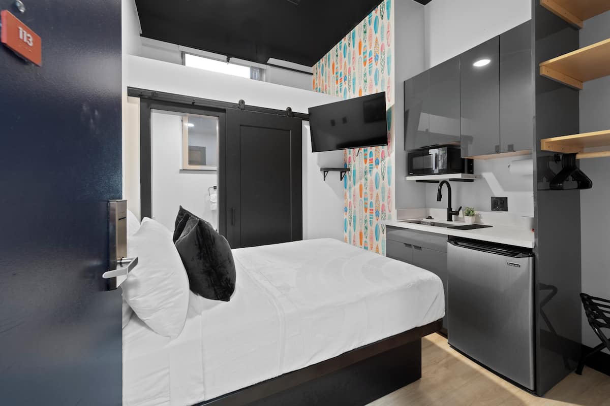 Una habitación de hotel moderna con una cama, un pequeño rincón de cocina, un televisor montado en la pared y un papel tapiz decorativo.