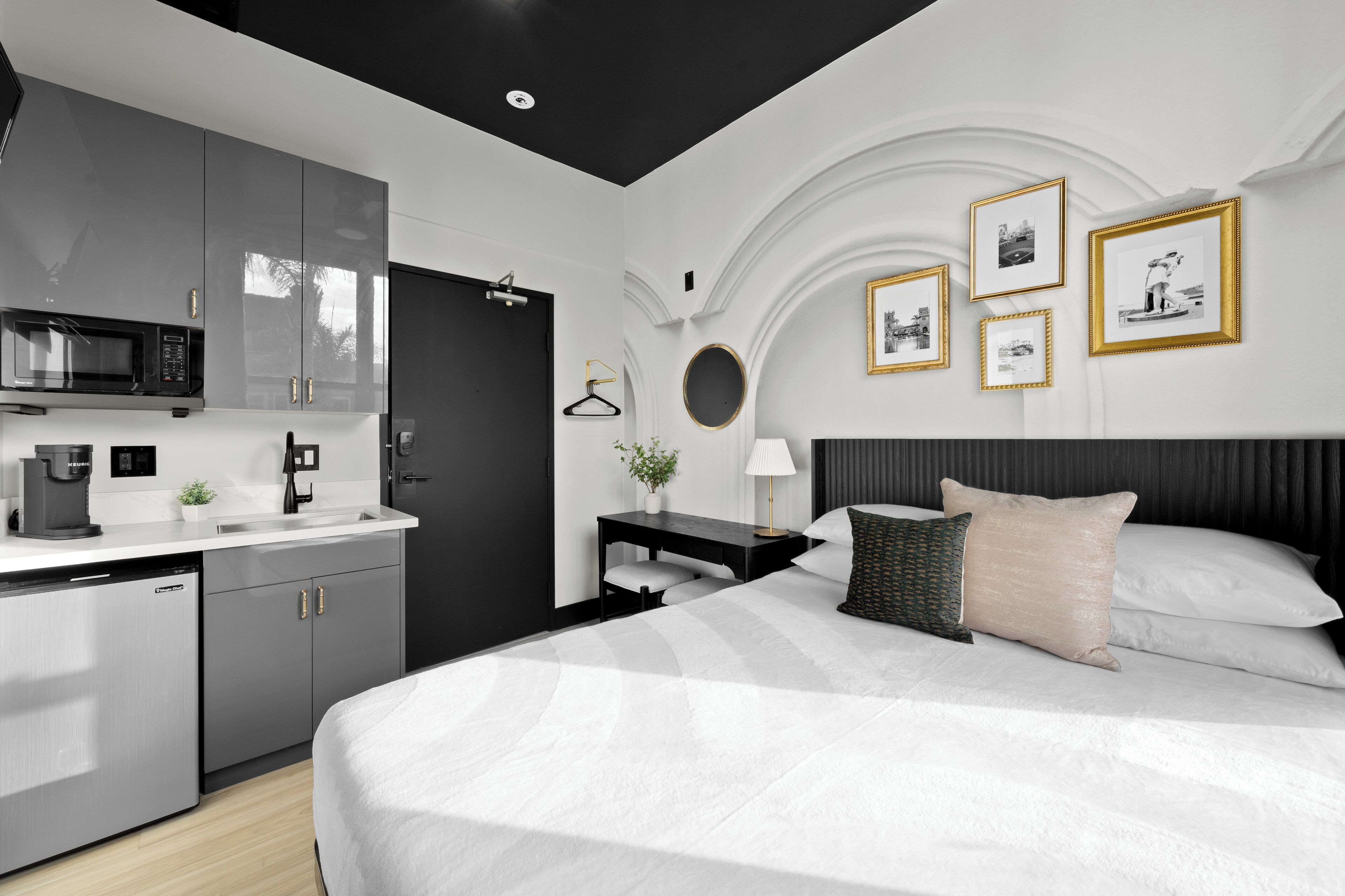A modern hotel room with a large bed, a bedside table with a lamp, a small desk with a chair, a microwave, and a coffee maker.