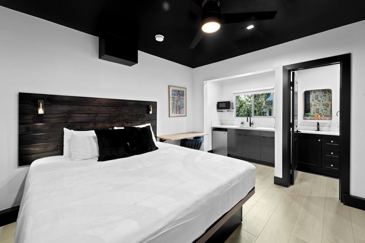 Un dormitorio moderno con una cama grande, un cabecero de madera oscura y una pequeña kitchenette al fondo.