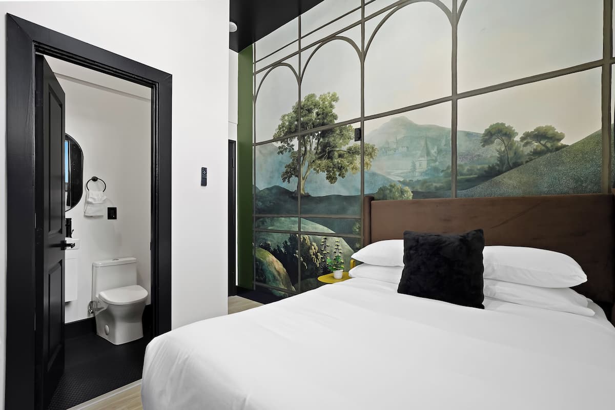 Habitación de hotel con cama, ventana con vistas a árboles y colinas, baño con inodoro y lavabo, y espejo.