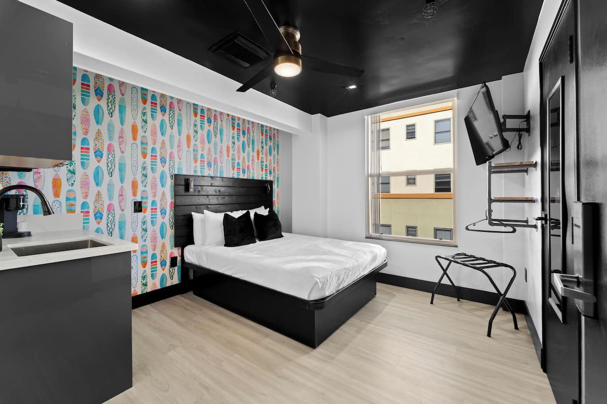 Un dormitorio moderno con cabecera negra, una cama grande, una pequeña cocina con pileta y una ventana con vista a edificios.