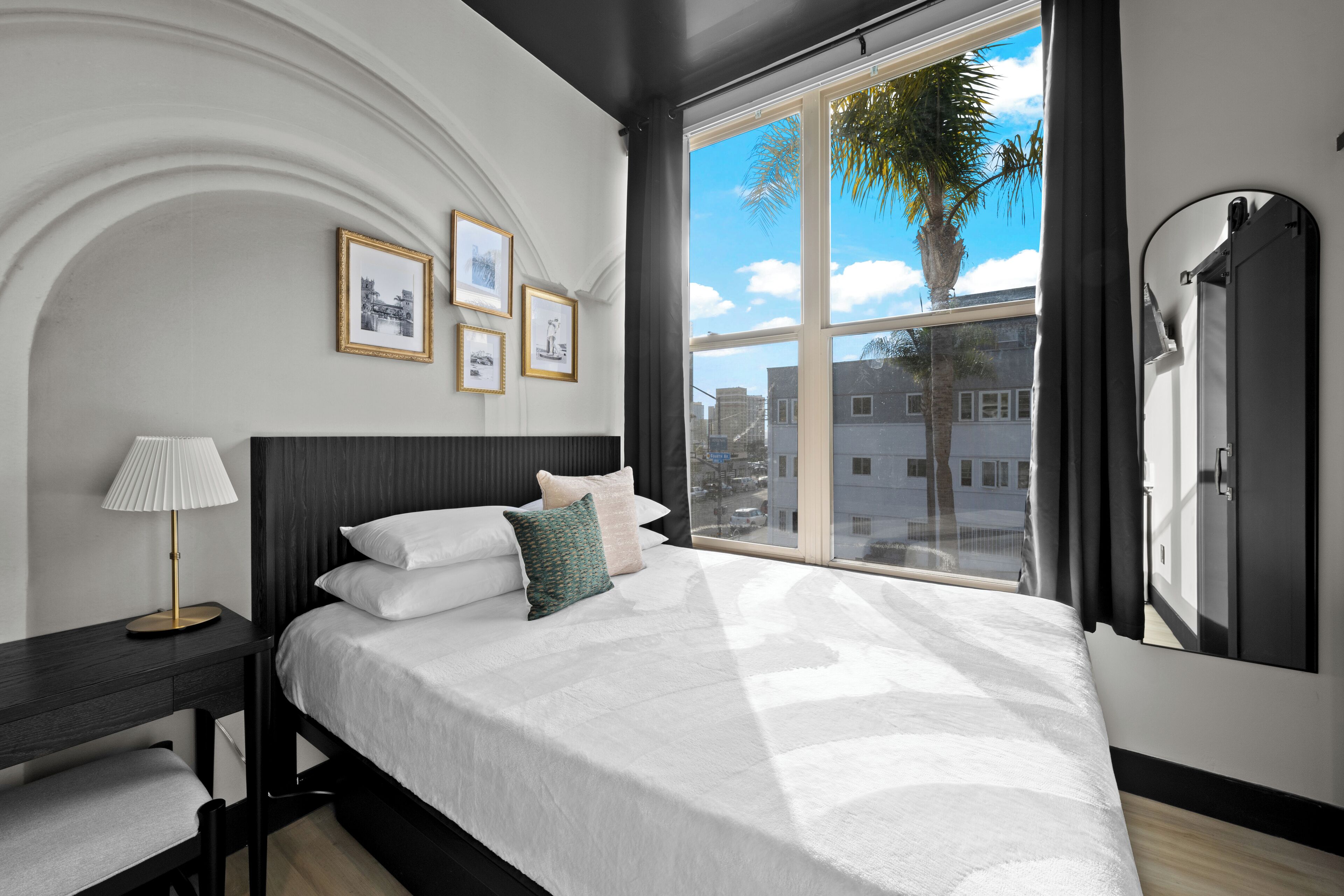 A modern bedroom with a large bed, a bedside table with a lamp, a large window with a view of buildings and a palm tree, and framed pictures on the wall.