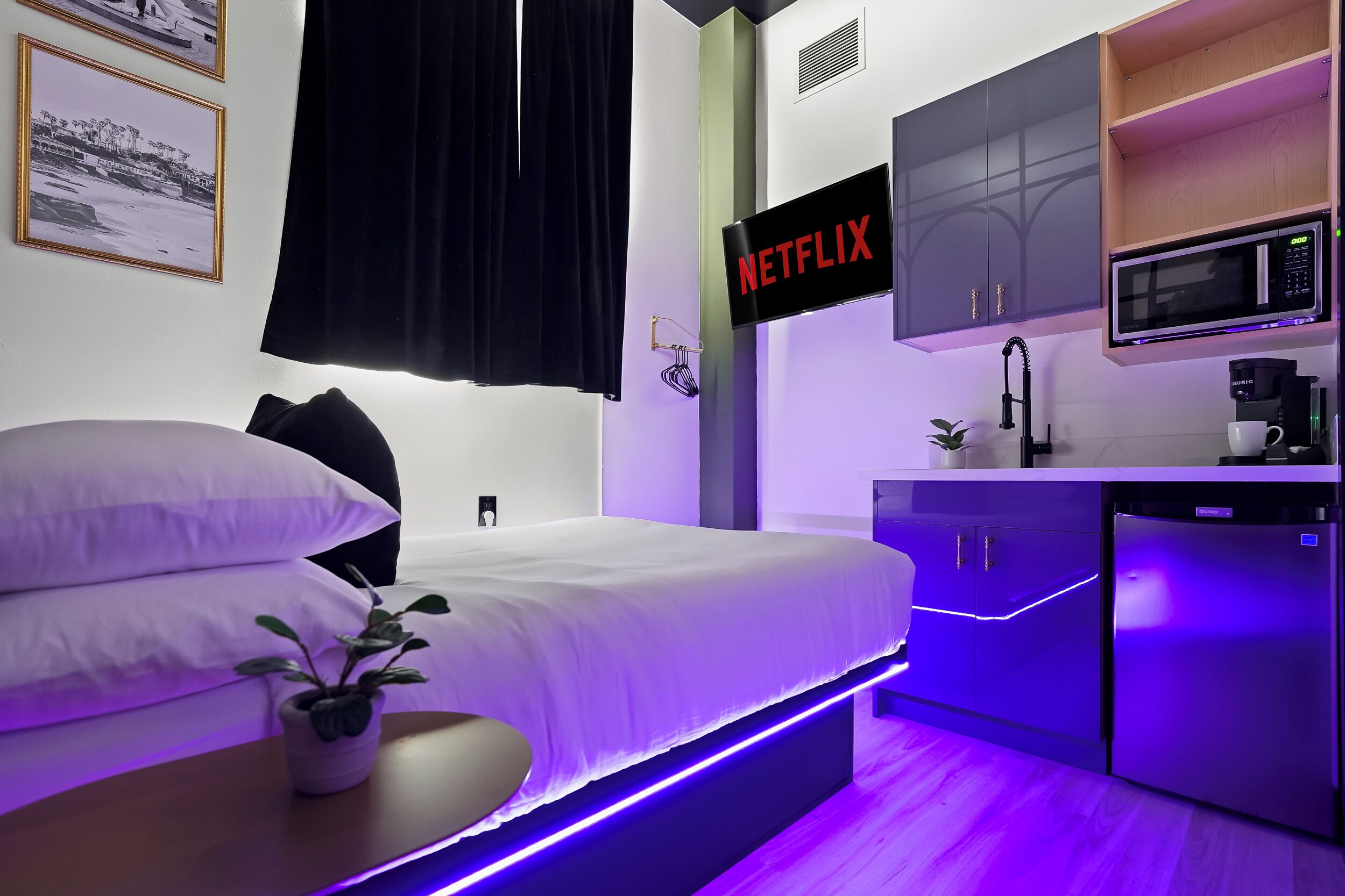 Una habitación de hotel moderna con cama, planta en la mesita de noche, cartel de Netflix en la pared, microondas y cafetera.