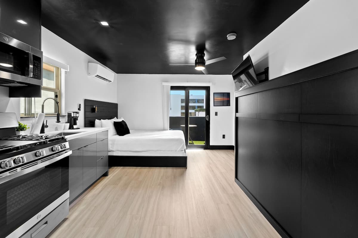 Una cocina moderna con estufa integrada, microondas y lavabo. Se ve una cama con cabecera negra y un ventanal grande al fondo.
