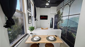 Un espacio habitable compacto con una mesa de comedor preparada para dos personas, un sofá, un televisor instalado en la pared y un ventanal con cortinas.