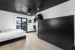 A modern bedroom with a bed, a large black headboard, a flat-screen TV, and a ceiling fan. - The Neon Elm Hotel (San Diego)
