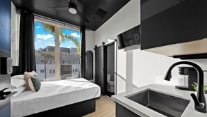 A modern hotel room with a bed, a large window with a view, a kitchen area with a sink and stove, and a ceiling fan. - The Neon Elm Hotel (San Diego)