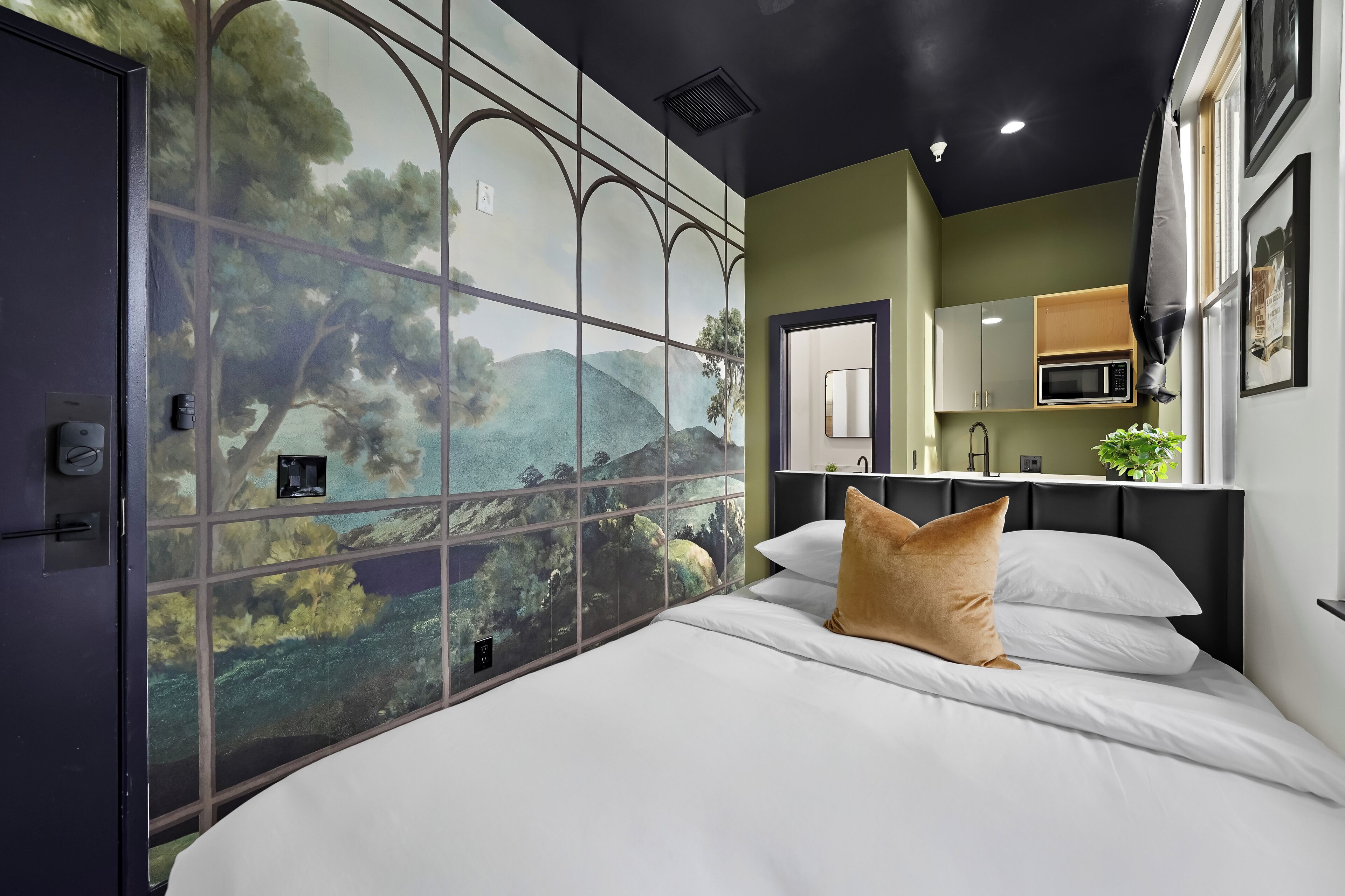 Ein Schlafzimmer mit großem Fenster, einem Bett mit weißer Bettwäsche und einem Kissen, einem Nachttisch und Blick auf einen Küchenbereich.