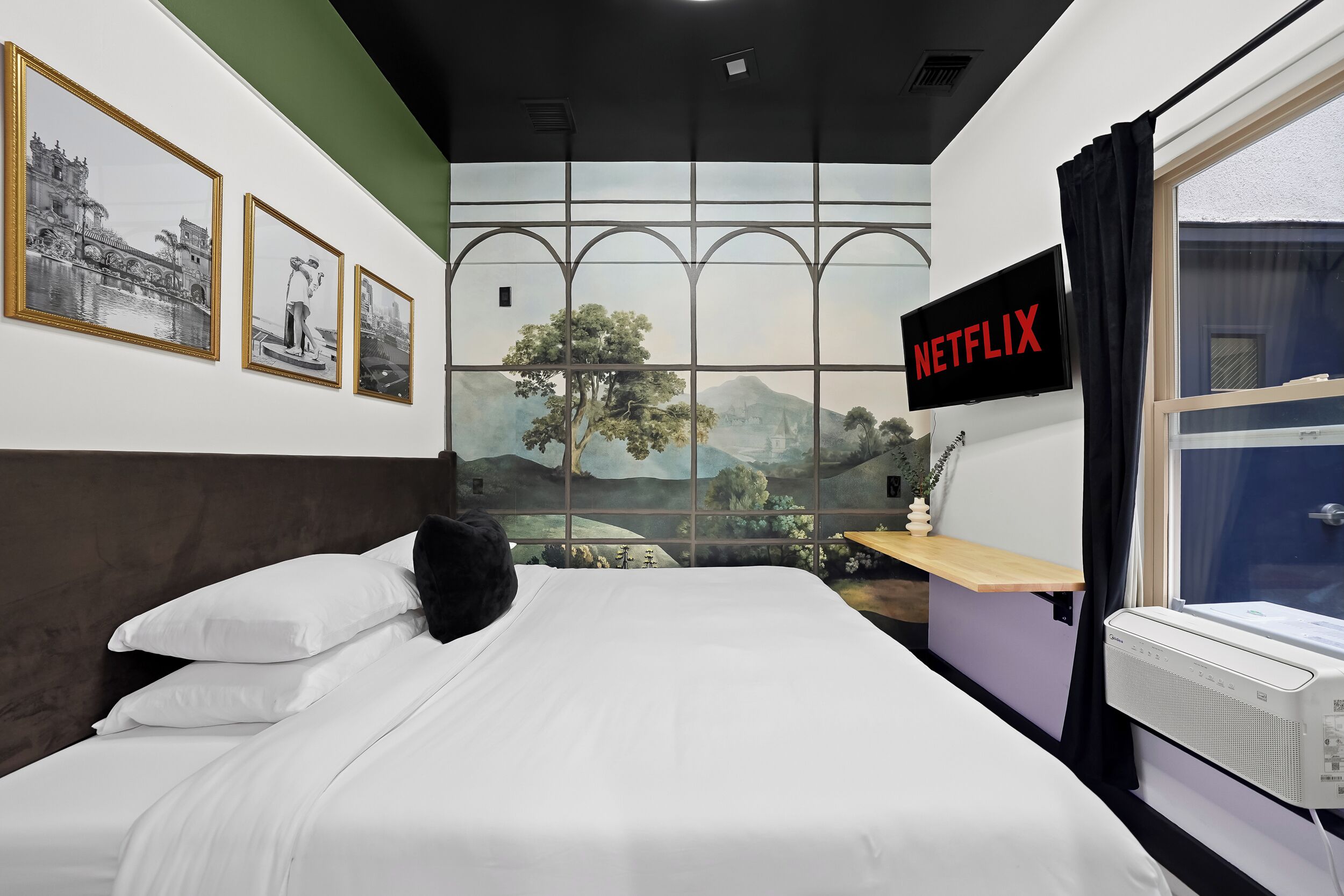 A hotel room with a bed, a wall-mounted TV, a window with curtains, and framed pictures on the wall.