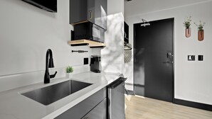 A modern kitchen with a black refrigerator, a built-in microwave, and a sink. There is a wooden shelf above the microwave and a small plant on the counter. - The Neon Elm Hotel (San Diego)