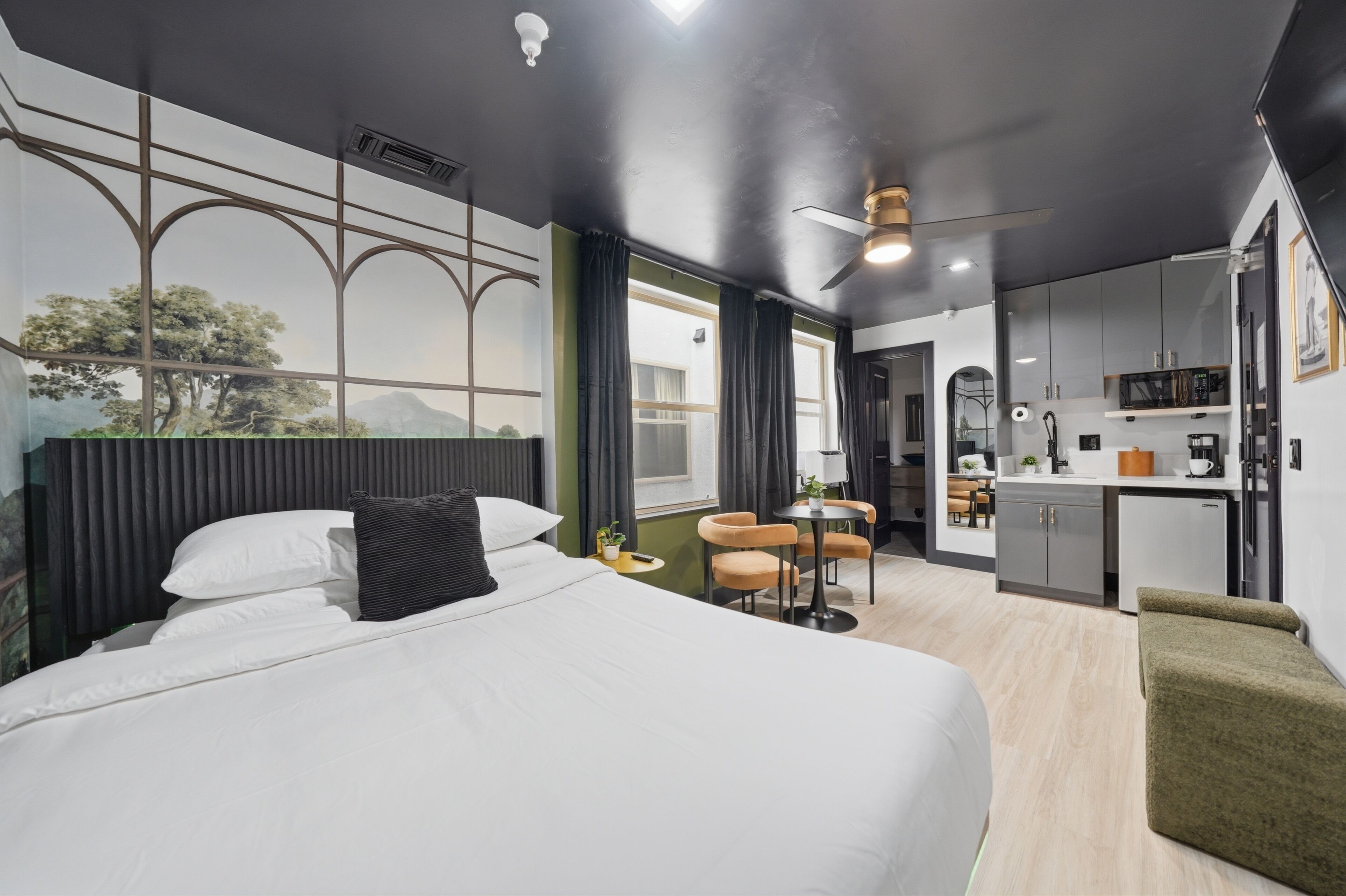 Une chambre d’hôtel moderne équipée d’un lit, d’un bureau, d’une chaise, d’une petite kitchenette et offrant une vue sur les arbres par la fenêtre.