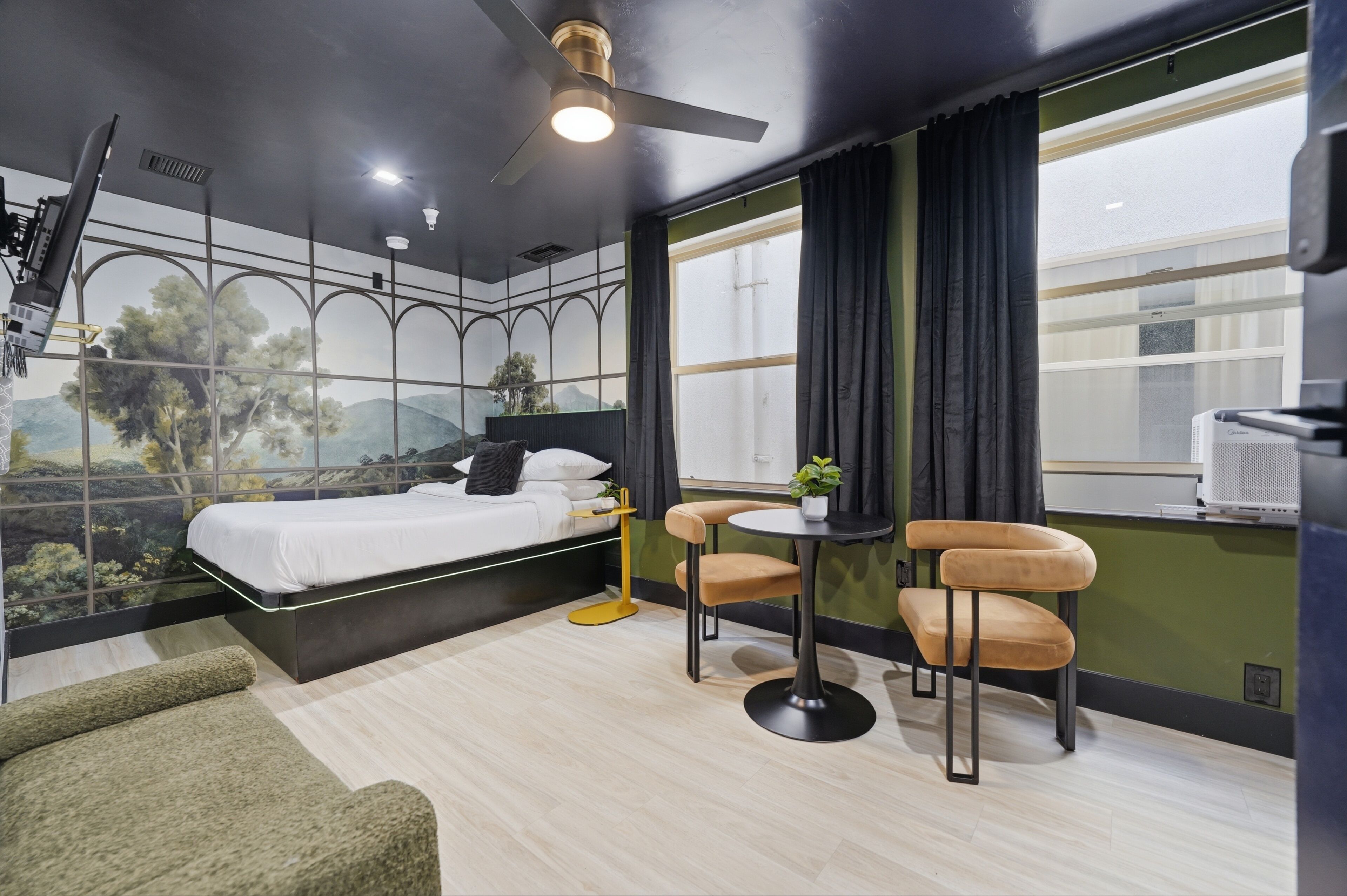 Una habitación moderna con cama, escritorio, dos sillas, ventilador de techo y un ventanal con cortinas.