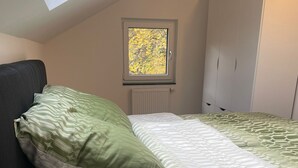 Una camera da letto con soffitto spiovente, una finestra vista alberi, un letto con biancheria verde e bianca e un armadio bianco.