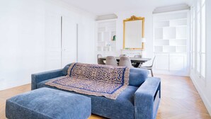 A spacious living room with a blue sofa and ottoman, a dining table with chairs, and a large mirror with a gold frame. - Charming 1-Bedroom by Champ de Mars (Paris)