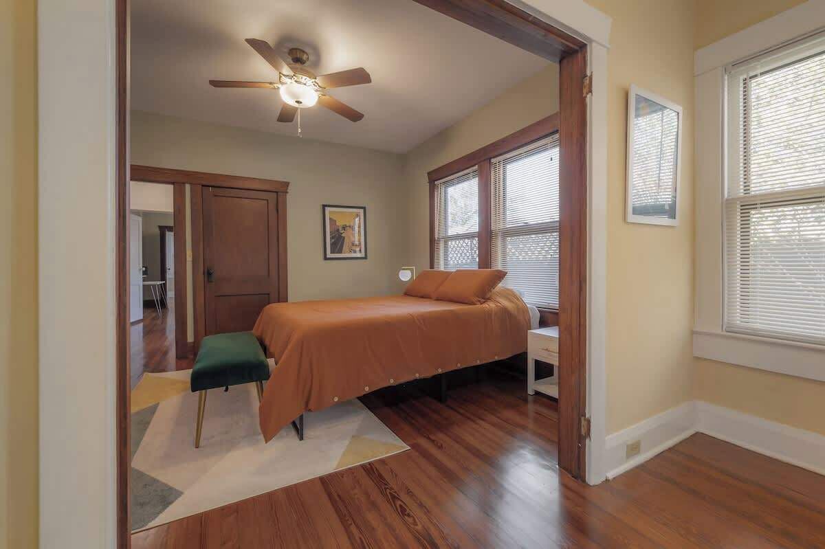 Un dormitorio con cama, ventilador de techo, ventana con persianas y puerta que da a otra habitación.