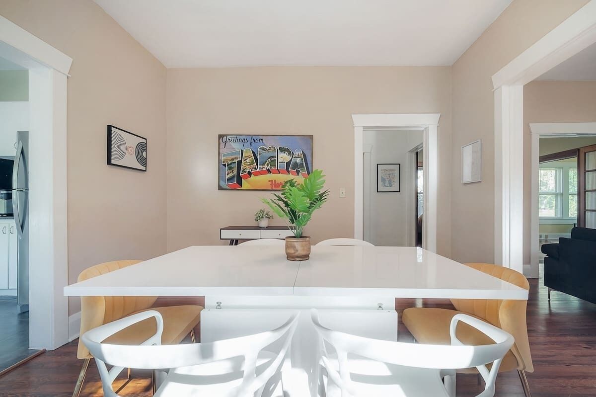 A dining area with a large white table, four chairs, a potted plant, and a wall with a framed picture and a painting.