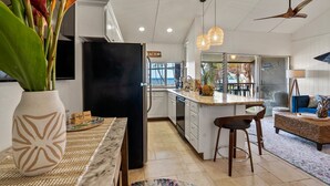 A modern kitchen with white cabinets, a black refrigerator, and a dining area with a bar stool and a woven coffee table. - Oceanfront Stunner w/ Cruiser Bikes & Pickleball (Kihei)