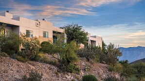 A modern building with a flat roof and large windows, surrounded by desert vegetation and mountains in the background. - Starr Pass Spring Golf Retreat - April 12-15 - 1 Bedroom Condo (Tucson)