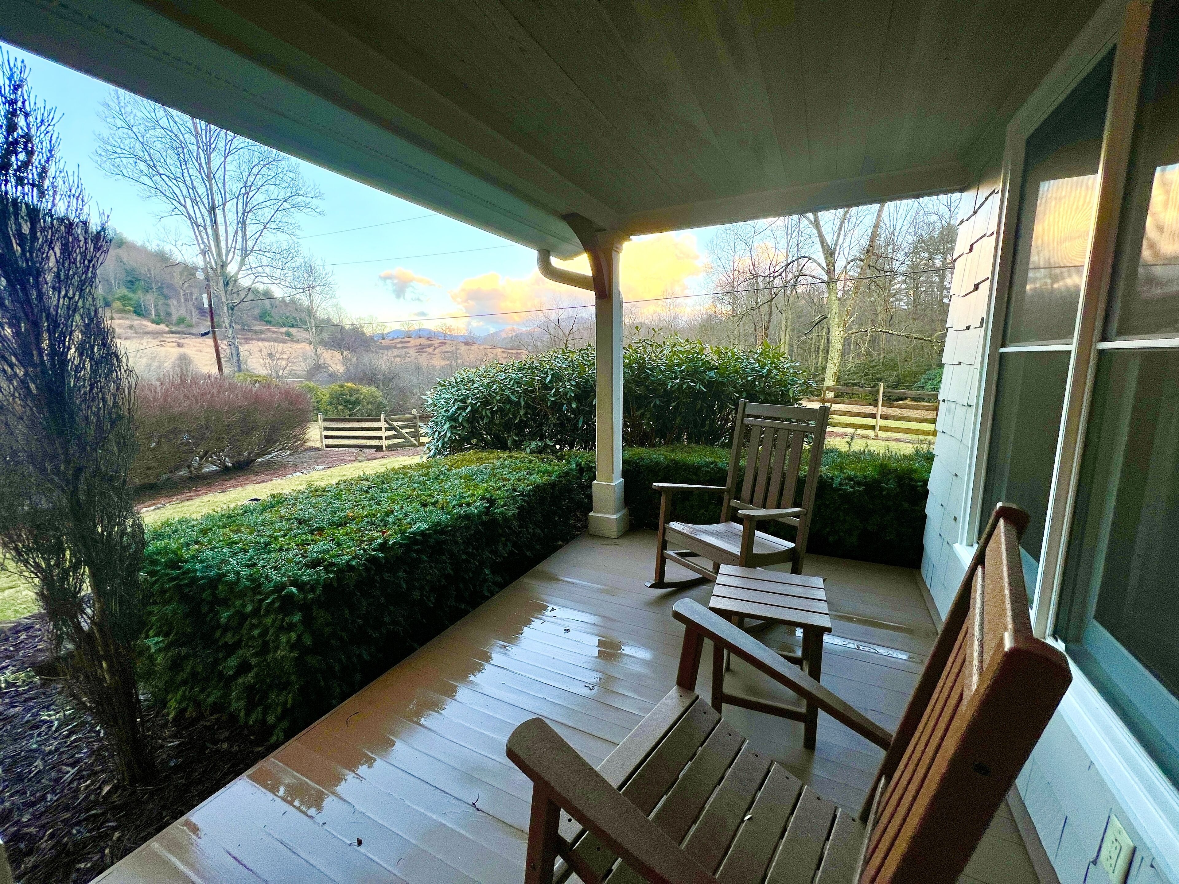 Eine hölzerne Veranda mit einer Bank und einem Sessel, mit Blick auf einen gepflegten Garten und eine weite Landschaft.