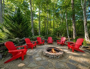 A circular fire pit surrounded by six red Adirondack chairs in a forest setting.
